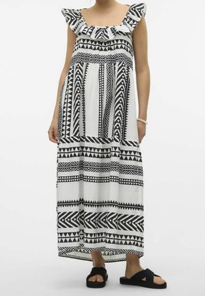 Femme portant une robe maxi à motif géométrique noir et blanc avec un col volanté et des sandales noires à enfiler.