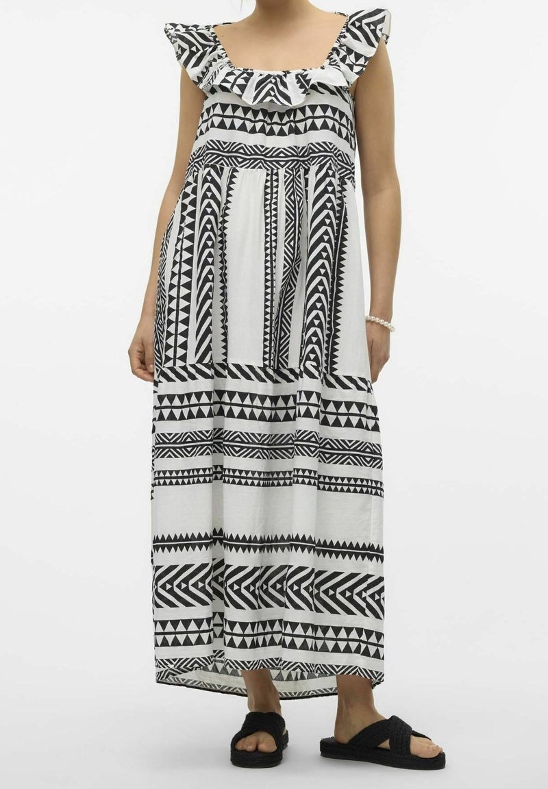 Femme portant une robe maxi à motif géométrique noir et blanc avec un col volanté et des sandales noires à enfiler.