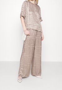 Completo due pezzi beige con motivo in tessuto leggero; la parte superiore presenta maniche corte e una vestibilità ampia, abbinata a pantaloni a gamba larga. Borsa bianca e scarpe.