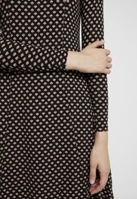 Kvinna i en svart klänning med små geometriska mönster, med armarna korsade, som fokuserar på ärm- och handdetaljer.