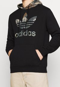 Svart sweatshirt med en huva i kamouflagemönster och en stor grå "adidas"-logotyp. Har en framficka samt ribbade muddar och fåll.