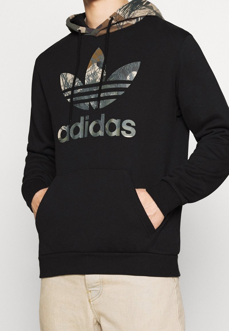 Svart sweatshirt med en huva i kamouflagemönster och en stor grå "adidas"-logotyp. Har en framficka samt ribbade muddar och fåll.