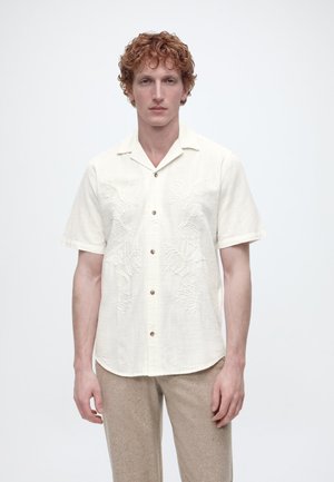 Mężczyzna o kręconych rudych włosach nosi kremową koszulę z krótkim rękawem z kwiatową haftem oraz jasnobeżowe spodnie na jednolitym białym tle.