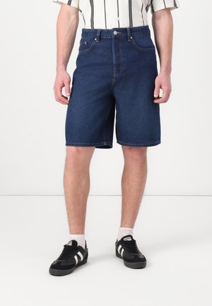 Shorts en denim foncé à taille haute, avec des poches avant, une fermeture à bouton et une coupe droite. Portés avec des baskets noires et des chaussettes blanches.