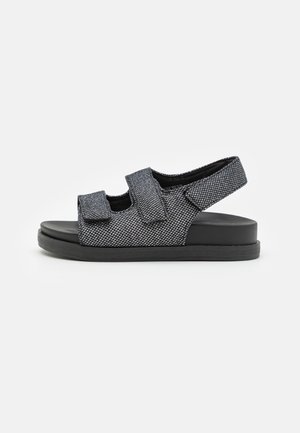 Sort og grå tekstureret sandal med åben tå med to justerbare remme over foden og en rundt om hælen, på en flad sål.