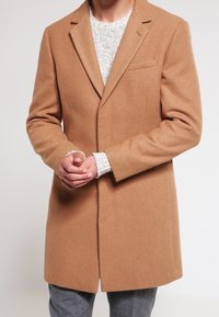 Camel uldbland frakke med skræddersyet design, notched lapels og sidelommer, båret over en let struktureret sweater og grå bukser.