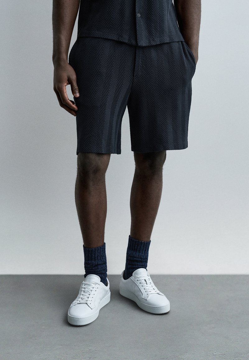 Partie inférieure du corps d'une personne portant un short texturé foncé, des chaussettes bleu marine et des baskets blanches, debout sur un sol gris devant un arrière-plan uni.