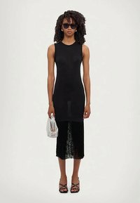 Femme en robe noire sans manches avec un ourlet frangé, talons noirs à lanières, lunettes de soleil, tenant une pochette texturée blanche devant un fond blanc.