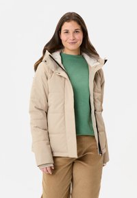 Beige gewatteerde jas met capuchon, voorzien van een ritssluiting aan de voorkant, doorgestikt ontwerp en een zachte voering, gedragen over een groene trui en bruine broek.