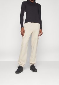 Person som bär beige cargojoggers med sidofickor, svart långärmad polotröja och svarta sneakers, står mot en enfärgad bakgrund.