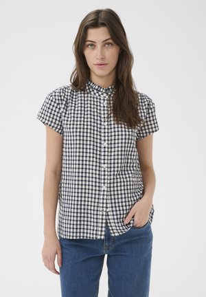 Jeune femme aux longs cheveux bruns portant une chemise à manches courtes à carreaux noirs et blancs et un jean bleu, debout avec une main dans la poche.