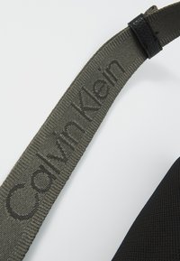 Melnā teksturētā auduma ar pelēku siksnu, uz kuras ir iepresēts "Calvin Klein" logo. Siksna ir ar gludu apdari un savienojas ar izturīgu melnu daļu.