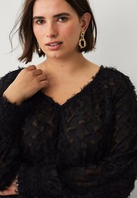 Kvinde med kort brunt hår iført store guldkædeøreringe og en sort tekstureret gennemsigtig bluse, mens hun holder sin krave med den ene hånd.