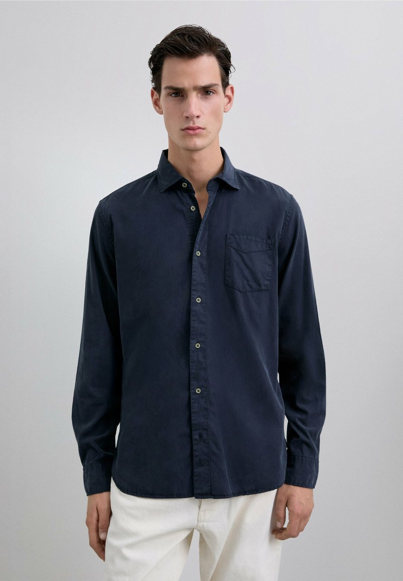 Camisa de manga larga azul marino en un tejido de algodón suave, con botones en la parte delantera, un solo bolsillo en el pecho y un escote relajado.