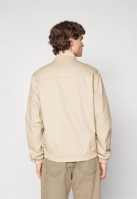 Beige jakke med opretstående krave, elastisk talje og manchetter, lavet af glat stof med minimalistisk design. Synlige sømme på ryggen.