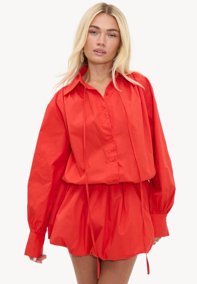 Femme blonde portant une chemise rouge vif à manches longues boutonnée avec un short ample assorti sur un fond clair uni.