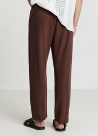 Braune, leichte Hosen mit lockerem Sitz, getragen von einer Person mit schwarzen Sandalen. Der Stoff hat eine glatte Textur und ein minimalistisches Design.