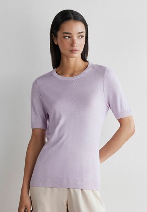 Kobieta w jasnofioletowej, krótkorękawkowej dzianinowej bluzce i beżowych spodniach, stojąca na tle jasnego, jednolitego tła, patrząca w bok.