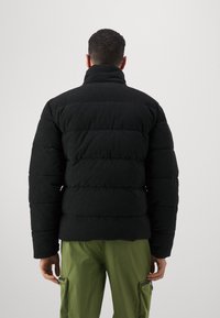 Schwarze Puffjacke mit geripptem Material, hoher Kragen und horizontalem Steppmuster. Kombiniert mit grünen Cargo-Hosen.