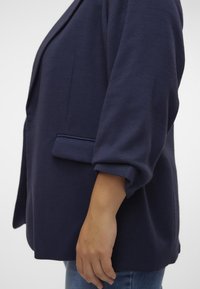 Navy blue blazer with textured fabric, single front pocket, rolled-up sleeves, and a tailored fit, worn over denim.
