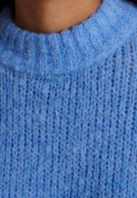 Gros plan sur une personne portant un pull en tricot épais bleu avec un col rond côtelé.