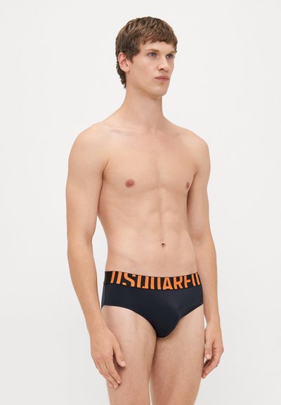 Oberkörperfreier Mann in schwarzen Badehosen mit breitem orangen Bund, auf dem große schwarze Schriftzüge zu sehen sind, vor einem schlichten weißen Hintergrund stehend.