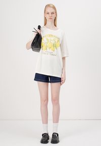 T-shirt oversize blanche avec un graphique de citron, associée à un short en jean noir, des chaussettes blanches et des chaussures noires à enfiler, portant un sac noir.