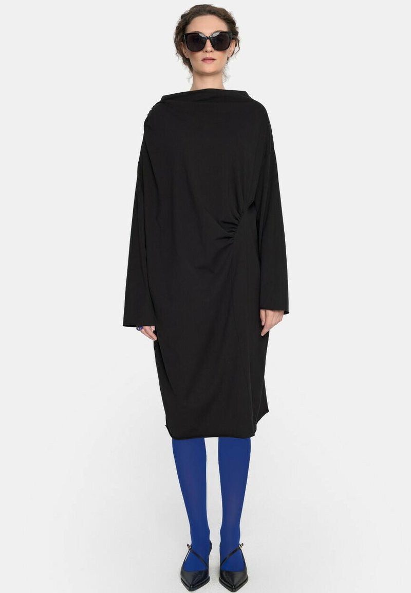 Czarna, oversize sukienka z długimi rękawami, opadającym dekoltem i detalem w formie skrętu po boku. Połączona z niebieskimi rajstopami i czarnymi, spiczastymi butami.