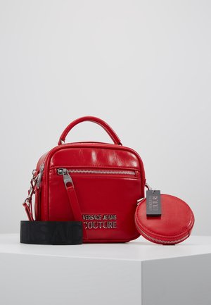 Rød lædertaske fra Versace Jeans Couture med top-håndtag, frontlomme med lynlås, aftagelig rem og matchende rund møntpose på hvid overflade.