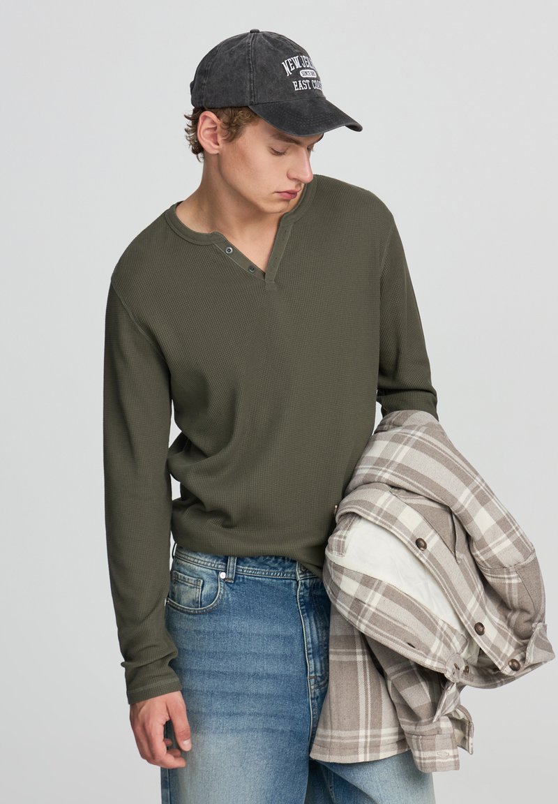 Maglia henley verde oliva in tessuto testurizzato, abbinata a jeans blu. Tiene in mano una camicia a quadri beige e bianca con bottoni e una fodera.