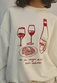 Sweatshirt crème avec des graphiques rouges de deux verres de vin, une bouteille et une assiette de pâtes avec une feuille. Texte en dessous dans une police de caractère ludique.