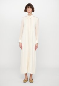 Vestido largo de crema claro con cuello, botonera y mangas largas transparentes; combinado con unos zapatos planos dorados y una textura suave.