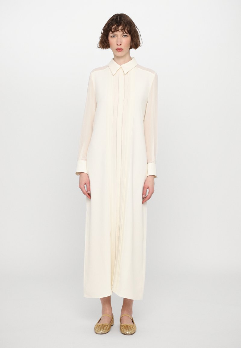 Vestido largo de crema claro con cuello, botonera y mangas largas transparentes; combinado con unos zapatos planos dorados y una textura suave.