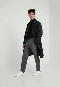 Zwarte lange jas over een zwarte coltrui, grijze geplooide broek en witte sneakers. Gladde stof, ontspannen pasvorm en ritsluiting op de jas.