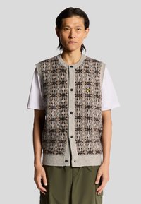 Gilet grigio lavorato a maglia con un motivo testurizzato marrone e beige, chiusura frontale con bottoni e un piccolo logo giallo sul lato sinistro del petto. Indossato sopra una maglietta bianca.