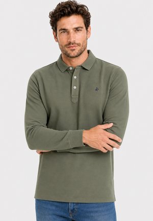 Hombre con cabello rizado que lleva una camisa polo de manga larga color verde oliva y vaqueros azules, de pie con los brazos cruzados.