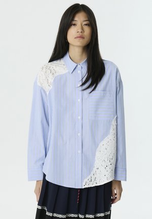 Camicia a maniche lunghe a righe blu e bianche con un pannello in pizzo su una spalla e una tasca sul petto. Tessuto strutturato, vestibilità comoda.