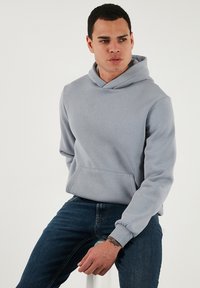 Világosszürke kapucnis felső, puha anyagból készült, elülső zsebbel, bordás mandzsettával és klasszikus húzózsinóros kapucnival, sötét farmernadrággal viselve.