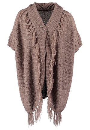 Femme portant un châle marron à franges, un haut blanc à manches longues, un jean bleu, des bottines noires, tenant un grand sac à main noir matelassé.