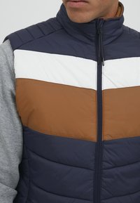 Pufový vest s vodorovnými pruhmi v námorníckej modrej, bielej a hnedej farbe. Zips vpredu, vysoký golier a prešívaná textúra. Viditeľné šedé rukávy.