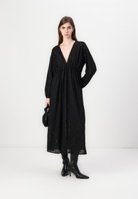 Μαύρο maxi φόρεμα από υφασμάτινη ύφανση, με βαθύ V-ντεκολτέ, μακριά μανίκια και ελαστική μέση. Συνδυάζεται με μαύρες μπότες μέχρι το γόνατο.