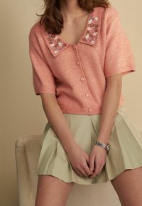 Różowy sweter z krótkim rękawem, ozdobiony haftem kwiatowym na kołnierzyku, zestawiony z jasnoszarym plisowanym spódnicą.