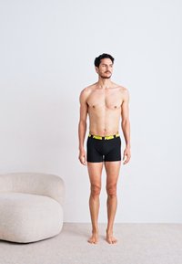 Boxeri negri cu o talie galbenă "PUMA", design ajustat, material moale, model stând pe un covor deschis într-un decor minimalist.