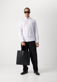 Bolso tote de cuero negro con asas dobles. El modelo lleva una camisa blanca de lino con botones y pantalones negros, acompañados de gafas de sol y zapatos negros.