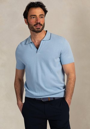 Man met donker haar en baard, draagt een lichtblauw poloshirt met korte mouwen en een donker broek met een patroonriem, handen in de zakken, kijkt naar rechts.