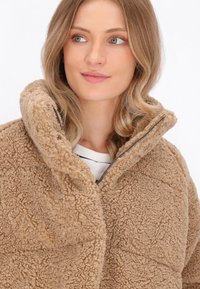 Beige, tekstureret fleecejakke med høj krave og lynlås. Blødt, fluffy materiale med et hyggeligt udseende.