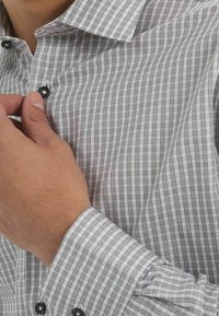 Chemise à carreaux gris et blanc avec un col à boutons, présentant une texture subtile et des boutons noirs. Main ajustant le col visible.