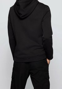 Černá mikina s kapucí a volným střihem, s žebrovanými manžetami a lemem, vyznačující se hladkou texturou. Nošena s korespondujícími černými kalhotami.