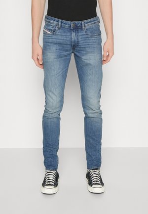 Wąskie, wyblakłe niebieskie dżinsy noszone przez osobę stojącą, zestawione z czarnymi niskimi trampkami i czarną bluzką.