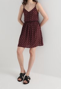 Robe florale bourgogne avec des bretelles spaghetti, des boutons décoratifs et une taille élastique. Associée à des sandales noires avec un accent à boucle ronde.